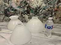 Three white vintage milk glass lamp shades displayed on a marble surface with a patterned background and a water bottle for scale.