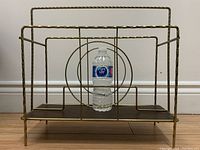 Front view of vintage MCM metal magazine/LP record rack with water bottle for scale, showing concentric circle design and wood base.