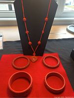 Full shot of cinnabar necklace on black stand with four red cinnabar bangles on red fabric.
