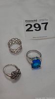Three silver rings laid out: floral patterned sterling silver ring, silver-toned ring with blue stone, and silver-toned ring with bow shape.