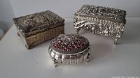 Three metal trinket boxes including one rectangular with floral relief, one square ornate with legs, and one round filigree silver box on red background.