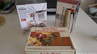Three boxed kitchen appliances on a table: Trudeau 3-in-1 fondue set, Stokes espresso and coffee maker, and metal apple peeler