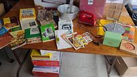 Wide view of the kitchen items on a table including vintage hand mixer, cookbooks, Bundy pan, towels, recipe cards, and metal pot.