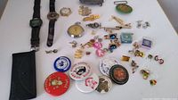 Overview of watches, button pins, tie clips, and lapel pins laid out on white surface showing quantity and variety