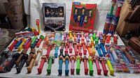 Wide view of large collection of Pez dispensers including boxed sets and various character dispensers arranged on surface.
