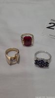 Three rings arranged on white surface showing red stone ring, clear stone ring, and blue cluster stone ring.