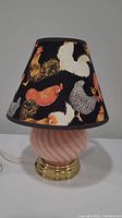 Full view of the table lamp showing the rooster-themed fabric shade on a black background and pink ceramic base with gold-tone bottom.