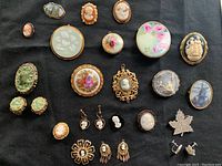 Top down overview of all brooches and earrings on black fabric background, showing variety of styles, shapes, and colors.