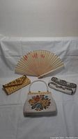 Full lot of three vintage beaded evening purses and one hand fan arranged on white cloth background.