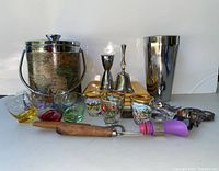 Photo showing ice bucket with world map design, stainless cocktail shaker, colored tilted shot glasses, decorated shot glasses, metal jiggers, snack plates, bottle stoppers and bar tool