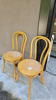 Two light wood bentwood chairs side by side, showing full front and side views, placed outside against a wall.
