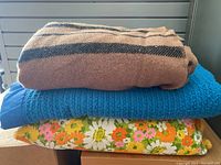 Three folded blankets stacked: a brown wool blanket with black stripes on top, a blue waffle blanket in the middle, and a floral colorful twin blanket with scalloped edges at the bottom.