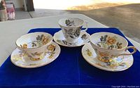 Three teacups with matching saucers displayed on a blue cloth, showing floral patterns of yellow and grey roses with gold trim.