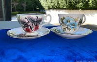 Two Queen Anne china teacups and saucers on a blue cloth, each cup with distinct floral patterns and gold trim.