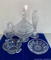 Five crystal items on blue cloth: tall vase, lidded candy bowl, crystal basket, silver overlay bowl, and small round dish.