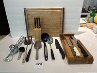 Photo showing assortment of kitchen tools including cutting boards, knives in block, various spatulas, ladle, serving spoon, and handheld egg beater mixer.