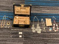 Jewelry collection displayed with jewelry box and multiple necklaces, earrings, pendants, and rings spread on a carpeted floor.