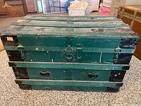 Front view of vintage green and black steamer trunk on carpeted floor.