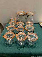 Twelve clear glass storage jars arranged on a table in rows, showing metal clamp closures and orange rubber seals.