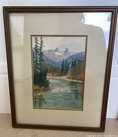 Full view of the framed watercolor painting showing a river, trees, and mountain peaks with beige matting and wood frame.
