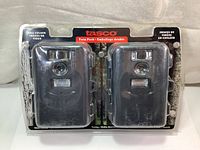 Front view of the new sealed Tasco trail camera twin pack, showing two black cameras in plastic blister pack with label detailing features.