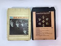 Two 8-track tape cartridges: Meet The Beatles! and Jesus Christ Superstar. The Beatles tape is cream-colored with their photo on the label; Jesus Christ Superstar tape is black with a brown label showing theatrical artwork and song listings.