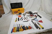 Overview of the tool selection arranged on a white surface, showing multiple screwdrivers, pliers, a hammer, scissors, utility knives, tape measure, and electric drill.