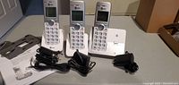 Three white and grey cordless handsets on base stations with charging cords and user manual on a table.