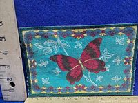 Front side of antique cigarette felt showing a red butterfly with a floral border on blue-green felt with ruler indicating size about 5 inches wide.