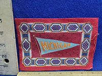 Front view of the red felt with geometric border and Michigan pennant in the center, with ruler for scale showing approx. 3x2 inches.