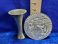 Two brass items shown: a circular brass tile with raised dragon motif about 3 inches diameter, and a small fluted brass vase about 6 inches tall.