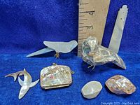 Photo showing assorted abalone and mother of pearl items: carved birds including one clear mother of pearl bird with etched wing details and gold metal legs, an abalone shell bird with metal claw feet, a small abalone shell change purse, and two iridescent shell beads placed before a ruler for sizing reference.