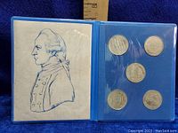 Blue presentation book open to a page displaying five commemorative silver dollars and a sketch of Captain Cook.