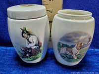 Two vintage pottery jars with artwork; one has lid with lion emblem and girl image, other has dog image without lid.