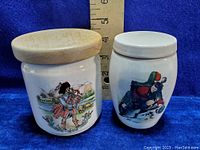 Two vintage pottery jars showing front Scottish illustrations and wooden and ceramic lids, set against a blue fabric background with a ruler showing approx 4.25 inch height