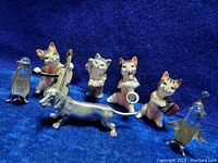 Seven animal figurines arranged on blue fabric. Includes three cat figurines playing musical instruments, a dachshund dog, glass penguin and seal figurines.