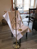 Full view of tall brass towel rack with four rings and smaller washcloth rack draped with white cloth on chair and floor, visible water and rust spots on base of tall rack