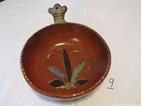 View of entire bowl showing handle shaped like a bird or animal head and painted leaf design inside bowl on reddish-brown base.