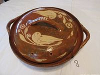 Top view of brown ceramic covered casserole dish lid with white bird and floral designs and handle, showing holes and handle repair.