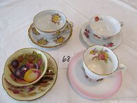 Four bone china teacups and matching saucers displayed together on a cloth surface, showing different floral and fruit hand-painted designs and gold trim.