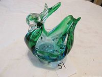 Green and clear glass art figurine in shape of a swan with detailed head and curved neck, shown from front angle on a white cloth surface.