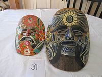 Photo showing both Mexican clay masks on a table, the larger one mostly black with floral designs, the smaller one colorful with prominent floral decorations and a mustache.