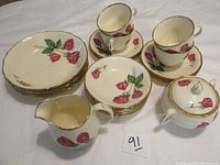 Full set including teacups, saucers, creamer, sugar bowl, fruit and dessert bowls with rose floral pattern and gold trim scattered on table.