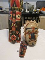 Three hand painted wooden masks displayed upright against a white wall and kitchen background. The large mask has bold red and black geometric designs; the medium mask is pale with raised features; the small mask shows colorful dot patterns.