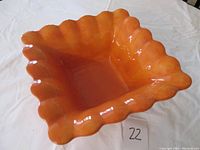 Top-down view of the orange scalloped edge pottery bowl on a white cloth surface with a lot number card.