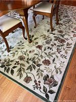 Close-up view of floral area rug with flowers and leaves in olive green, brown and earthy red on beige background, showing part of dining table and chairs on rug.