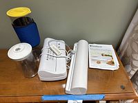 Photo showing FoodSaver Mini Plus vacuum sealer with two clear vacuum food containers, one with yellow lid and the other with white lid, and white vacuum bag roll holder.