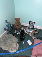 Photo showing various ceramic frogs, terracotta flower pot with lizard design, small decorative mirror, and green glass fish vase in corner on carpeted floor.