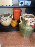 Green and cream-colored fall-themed ceramic vases with acorn and leaf accents, tall orange vase, and ceramic acorn container arranged on black shelving.
