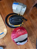 Three items on wooden floor: Coleman sleeping bag in red roll bag, Stinger wet/dry vacuum, white deck fan.